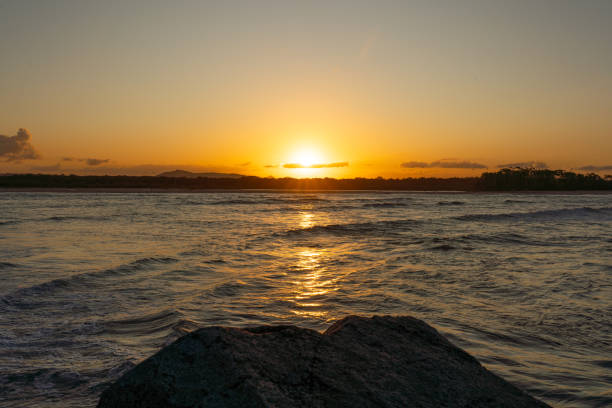 The sun is setting over the ocean, casting a warm glow on the water. The sky is a mix of orange and pink hues, creating a serene and peaceful atmosphere. The water is calm