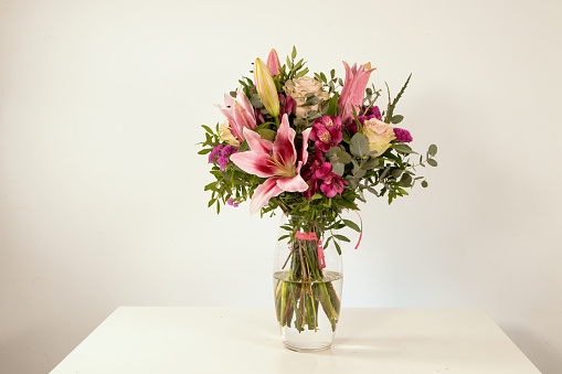 Bouquet of pink lilies with champagne-colored roses and decorative greens, on a white background and white wooden table