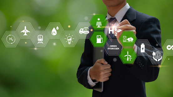 A businessman uses a virtual screen to illustrate green hydrogen production and usage, with icons representing solar panels, wind turbines, fuel cells, and hydrogen-powered vehicles. H2.