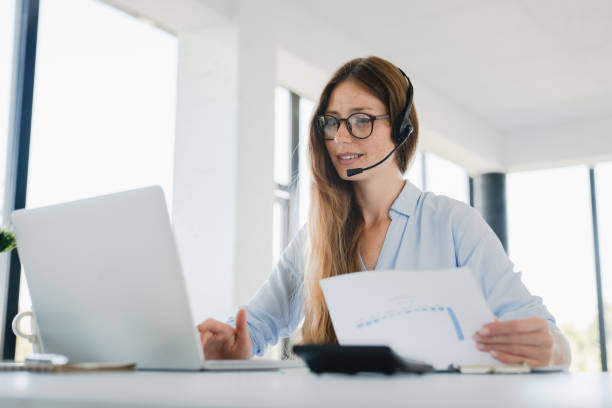 istock Young woman sales manager in headphones doing paperwork while on call with customer 2114991716