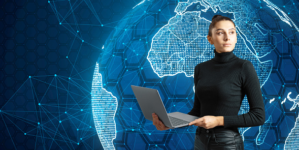 Attractive thoughtful young european businesswoman using laptop with glowing futuristic blue globe hologram on blurry background. Digital innovation and technology concept