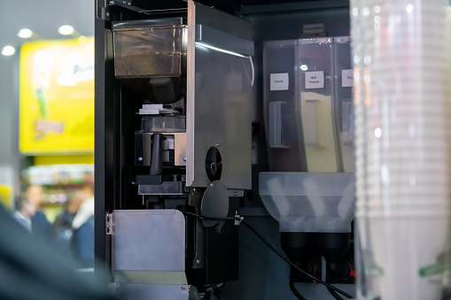 Close-up view of open bean-to-cup free standing vending coffee machine with grinder, brewer, soluble powder hoppers and paper coffee cup dispenser. Soft focus. Drink technology theme.