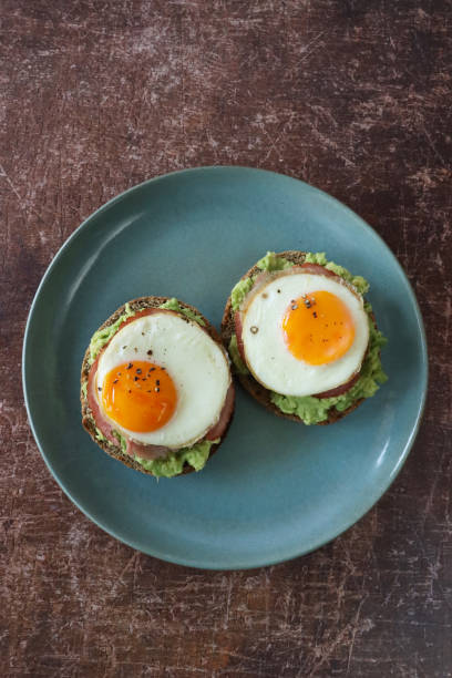 Image of breakfast, blue-green plate of wholemeal, toasted bagel topped with mashed avocado, ham bacon rasher, sunny-side-up fried eggs, cracked peppercorn seasoning, wood grain background, elevated view, focus on foreground