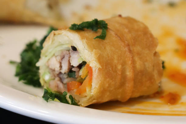 Stock photo showing close-up, elevated view of sliced pork and vegetable spring rolls, made with filo pastry and filled with a mixture of finely shredded vegetables including, spring onion, white cabbage, carrot, red pepper, bean sprouts and garlic, served on a white plate.