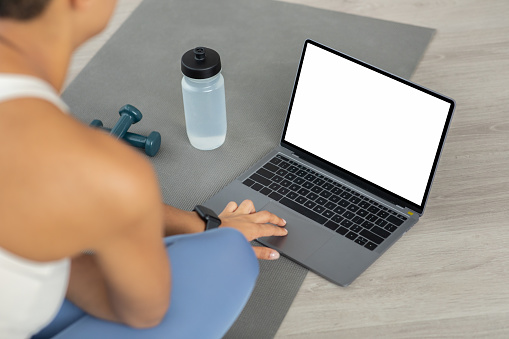 Over shoulder view of latin millennial woman using a laptop with a blank screen, surrounded by fitness equipment on a yoga mat, indicating a home workout planning or tracking