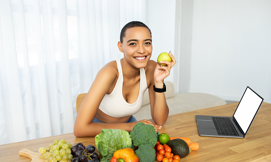 Cheerful latin woman holding green apple with variety of fresh fruits and vegetables on table, engaging in healthy lifestyle discussion over laptop with empty screen at home