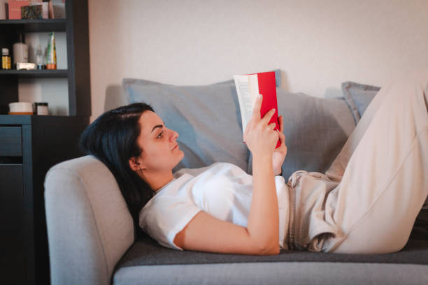 istock Beautiful young woman lying on her back and reading a book 2115098076