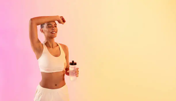 Relaxed fit latin millennial woman enjoying a break during her workout, holding a water bottle with eyes closed and a serene expression against a warm gradient background, studio