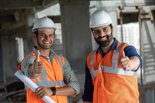 Portrait of confident male architects in protective workwear showing thumbs up signs while standing at construction site