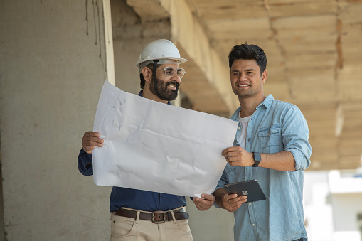 Smiling young man holding digital tablet and discussing blueprint with building contractor while standing at site