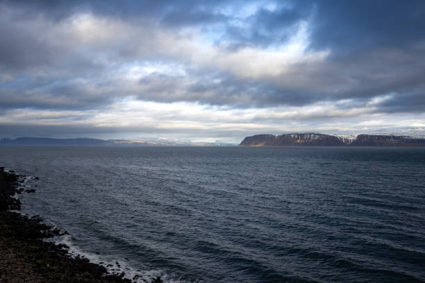 Calm water of Atlantic ocean. High mountains on the coast on the other side of fjord. Rocky coast in the foreground. Intense cloudy sky in the autumn. Area of Isafjordur, Westfjords, Iceland.