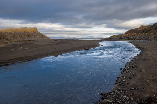 Dark volcanic sand beside the river flowing to the ocean. Mountains with cliffs on the horizon, with snow peaks. Area of Isafjordur, Westfjords, Iceland.