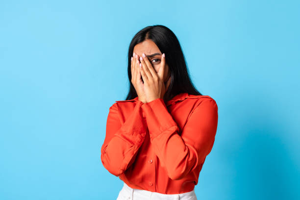 istock scared young indian woman peeking through fingers against blue backdrop 2115141885