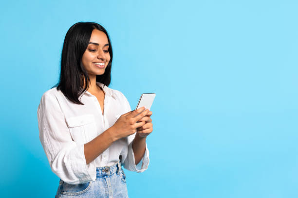 Cheerful middle eastern lady with smartphone texting over blue background Cheerful young middle eastern lady with smartphone texting and websurfing near copy space for mobile advertisement, standing over blue studio background. Communication, gadgets indian woman smiling stock pictures, royalty-free photos & images