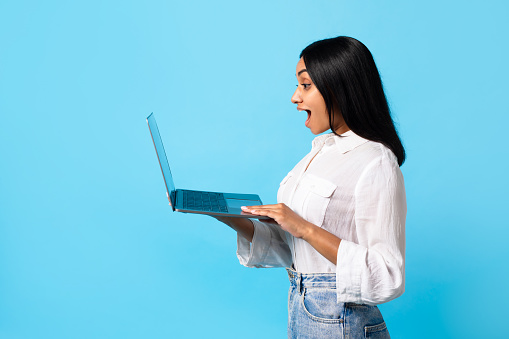 Online Work And Study. Side View Shot Of Excited Indian Woman Using Laptop And Looking At Computer Screen, Reading amazing News Online, Standing With Device On Blue Studio Background