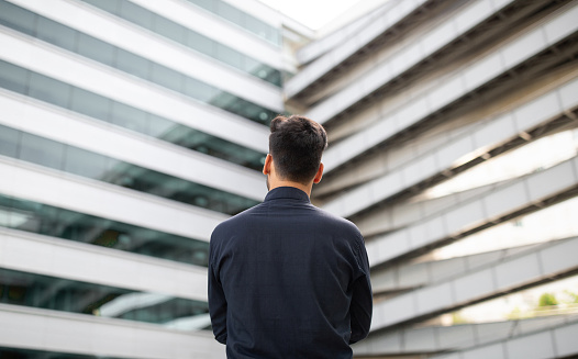 Unrecognizable young CEO businessman stands with his back to the camera outdoor, looking at modern office building, embodying the essence of success and corporate style. Copy space