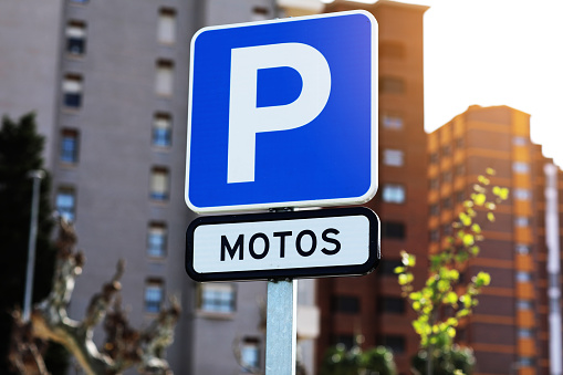 Blue sign with a big white P letter and inscription motos indicating the parking for motorcycles and scooters, on background residential buildings in the European city, Spain. Moto area.