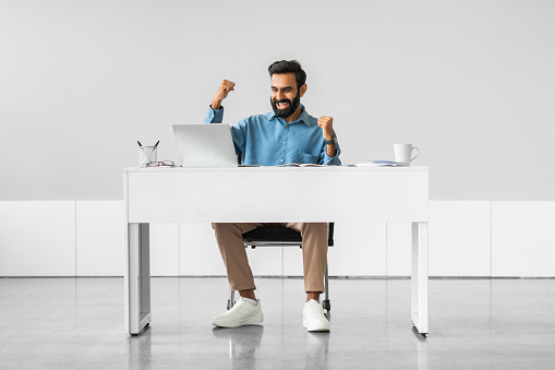 Elated indian professional man with raised arms showing jubilation at work success, seated at while modern desk with laptop and documents, full length