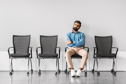 Confident indian businessman with a beard, sitting cross-armed in a vacant conference area, exuding a sense of authority and anticipation in a sleek, modern environment
