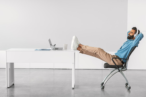 Content indian businessman reclining comfortably with his feet on the desk, reflecting moment of relaxation during busy workday in bright office, side view, free space