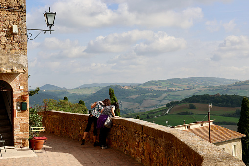 Pienza, Province of Siena, Tuscany, Italy - 25 October 2022: Two women survey the area. Pienza is a town and comune in the province of Siena, Tuscany, in the historical region of Val d'Orcia. The Val d'Orcia or Valdorcia is a region of Tuscany, central Italy, which extends from the hills south of Siena to Monte Amiata. Its gentle, cultivated hills are occasionally broken by gullies and by picturesque towns and villages such as Pienza, Radicofani and Montalcino. Its landscape has been depicted in works of art from Renaissance painting to modern photography.