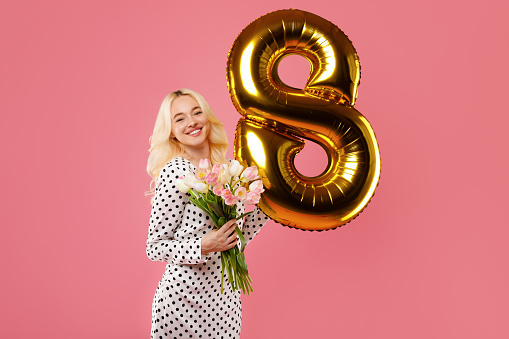 Radiant blonde woman holding bouquet of tulips and shiny golden balloon shaped as the number eight, embodying the spirit of International Women's Day