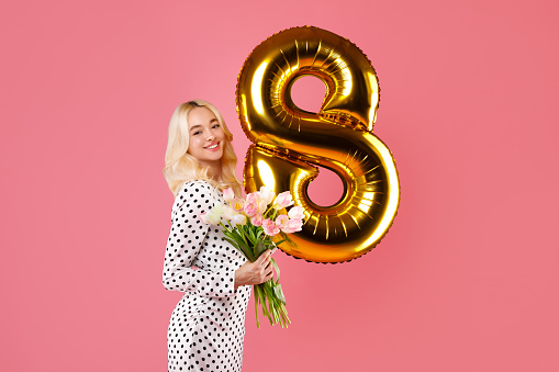 Smiling blonde woman in dotted dress holds pink tulips and golden number 8 balloon, celebrating Women's International Day on pink background