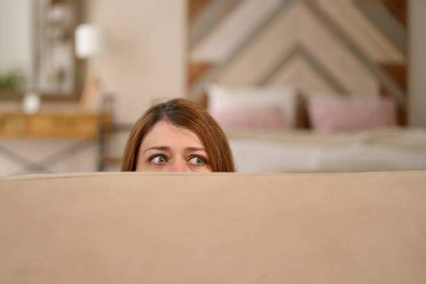 istock Young woman hides behind a sofa, notices surveillance 2115251309