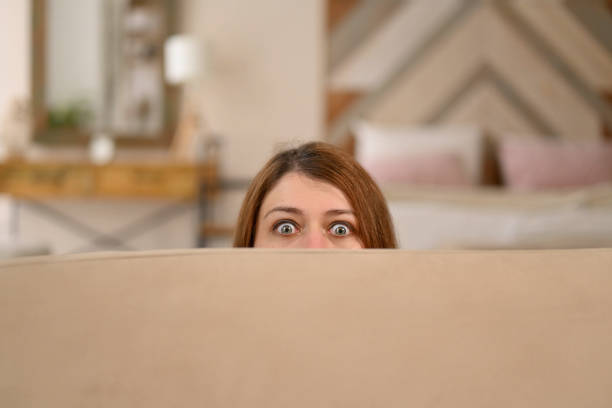 istock Young woman hides behind a sofa, notices surveillance 2115251311