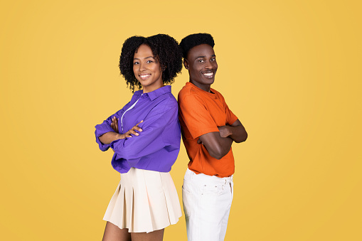 Confident young woman in a purple blouse and smiling man in an orange shirt, standing back-to-back with arms crossed against a bright yellow background, exuding positivity and teamwork