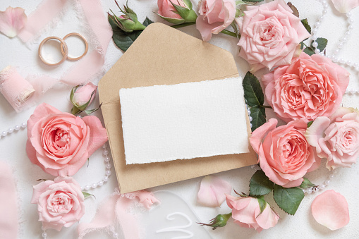 Blank card and envelope near light pink roses, wedding rings and silk ribbons on white table top view,  stationery mockup. Romantic flat lay with pastel decor  and flowers