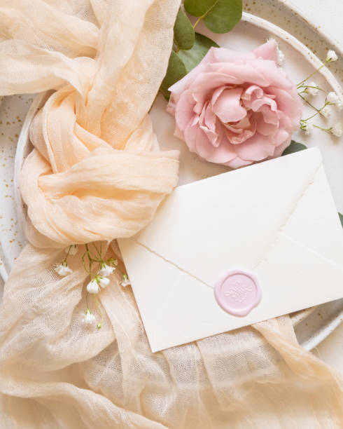 istock Envelope near cream fabric knot and light pink roses on plates top view copy space, wedding mockup 2115296871