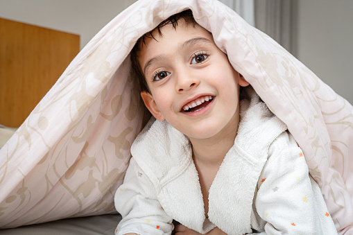 Smiling little boy hiding under blanket in bed after shower.