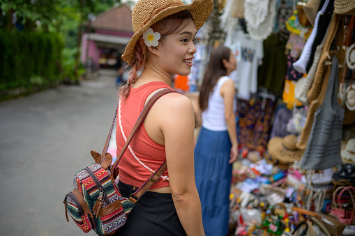A Young, Beautiful Asian Female Tourist Enjoys Street Shopping in Bali's Bustling Bazaar. Looking at camera.
