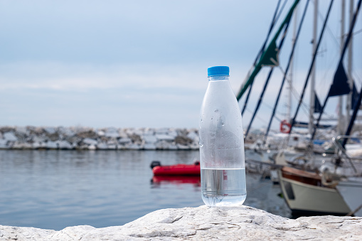 A transparent PET bottle half filled with water on a stone on the seashore. Sailboats are visible in the background. Symbolizing eco-awareness.