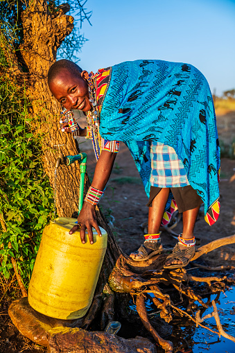 African woman from Maasai tribe collecting water to the tank, Kenya, Africa. When she finish filling the tank, she will carry water on her back to the village. African women and also children often walk long distances through the savanna to bring back containers of water. Some tourist camps cooperating with nearby villages and allow local people to use their water. Maasai tribe inhabiting southern Kenya and northern Tanzania, and they are related to the Samburu.