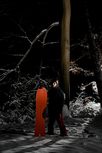 Young male snowboarder holding a splitboard looking into the distance while standing in the dark with low lighting on a snowy lawn near an old tree