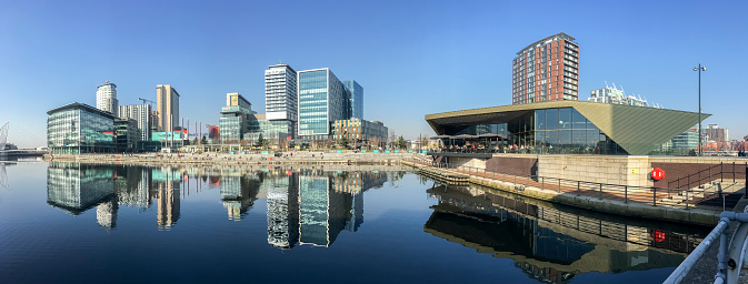 View across Salford Quays in Manchester UK.  Tall modern buildings can be seen. People can be seen on the promenade