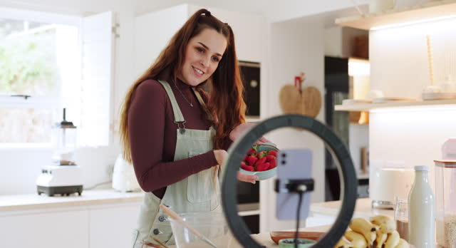 Woman, fruit and phone for live streaming food, cooking and meal for social media blog, vlog and breakfast. Waving hello, lighting or home influencer on technology for kitchen baking with strawberry