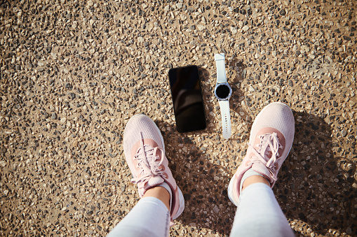Top view feet of a sportswoman in pink running sports shoes, a smart wrist watch and smartphone with black blank mockup digital screen on the asphalt. Copy ad space for sports and fitness mobile app