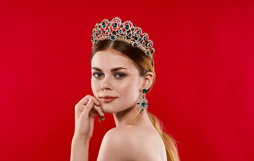 Portrait of a confident woman on a red background with a crown on her head and earrings on her ears. High quality photo