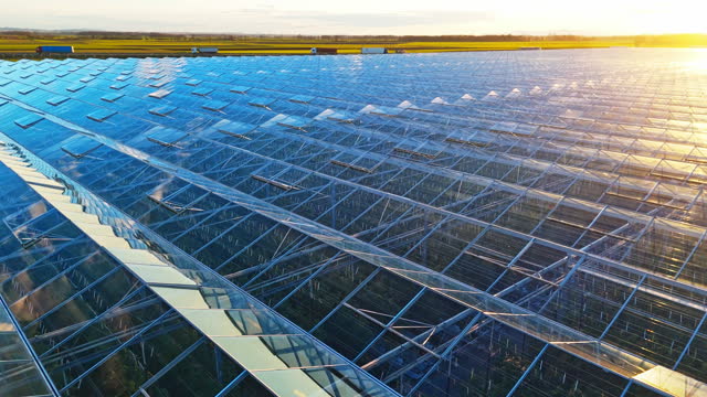 AERIAL Drone Speed Ramp Shot of Large Modern Glass Industrial Greenhouse Near Busy Highway Under Sky at Sunrise
