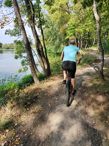 A young blonde woman rides a bicycle in the forest near the river on a path. Rear view. Tourism, sports and outdoor recreation