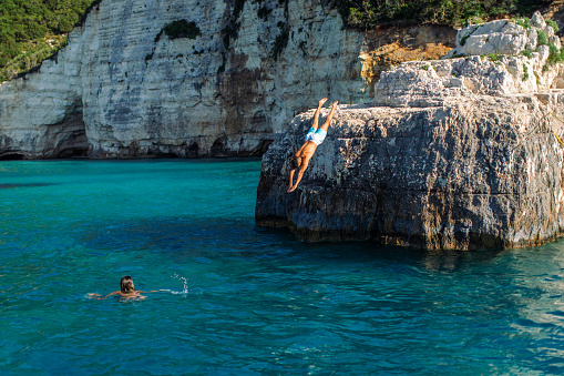 Handsome mid adult man jumping from the rock on coast and dives into the sea. One woman looking at him from the water