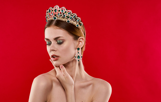 Portrait of a confident woman on a red background with a crown on her head and earrings on her ears. High quality photo