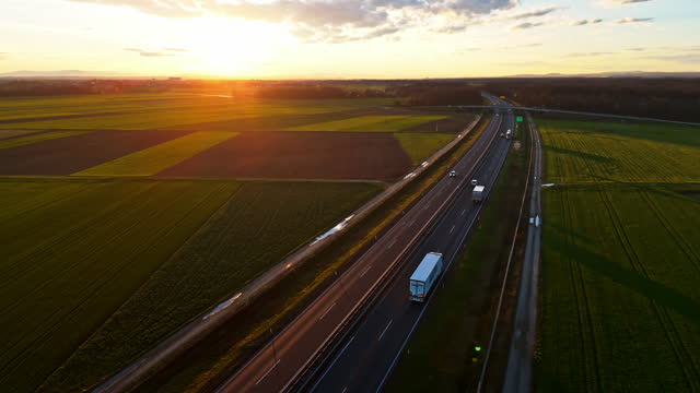 AERIAL Drone Flying Above Modern Glass Industrial Greenhouse Near Cargo Trucks Driving on Highway During Sunrise in Slovenia