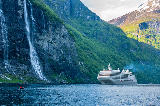 Powerful gigantic tree lined mountains and Seven Sisters waterfalls of Geirangerfjord dwarfing large ocean liner showing size perspective of the cliffs' height