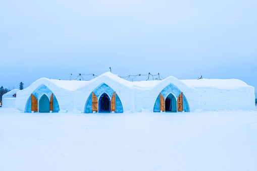 Full outside wide angle view facing the ice hotel and its three entrances made of blue ice and containing large brown wooden doors and no people in picture on a sub zero temperature day in the snow