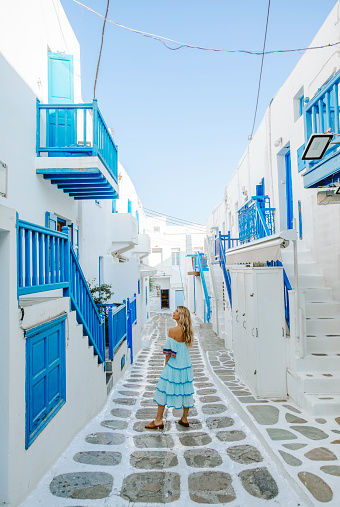 Rear view of a young beautiful happy traveler female lady in a blue dress enjoys looking up the scenery and walking through the traditional white and blue small, whitewashed alleys of Mikonos town, Cyclades islands of Greece during summertime.