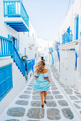 Rear view of a young beautiful happy traveler female lady in a blue dress enjoys the scenery and running through the traditional white and blue small, whitewashed alleys of Mikonos town, Cyclades islands of Greece during summertime.